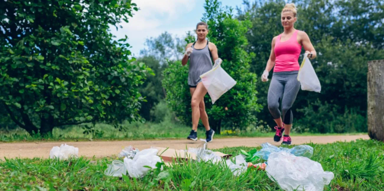 Two joggers carrying bags on a park path, with plastic waste in the grass in the foreground