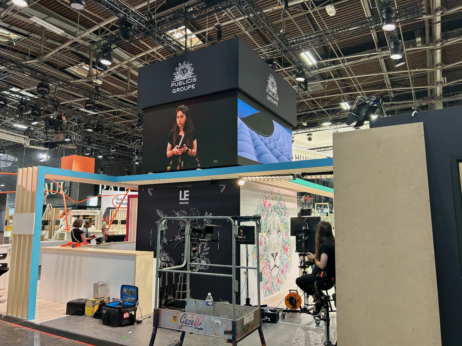 Publicis Groupe stand being assembled: LED screen cube suspended above the reception area, camera on a tripod in the foreground