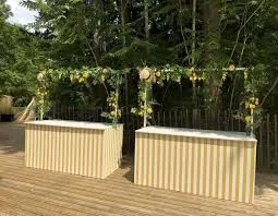 Two yellow and white striped counters decorated with plant garlands, on a wooden terrace surrounded by trees