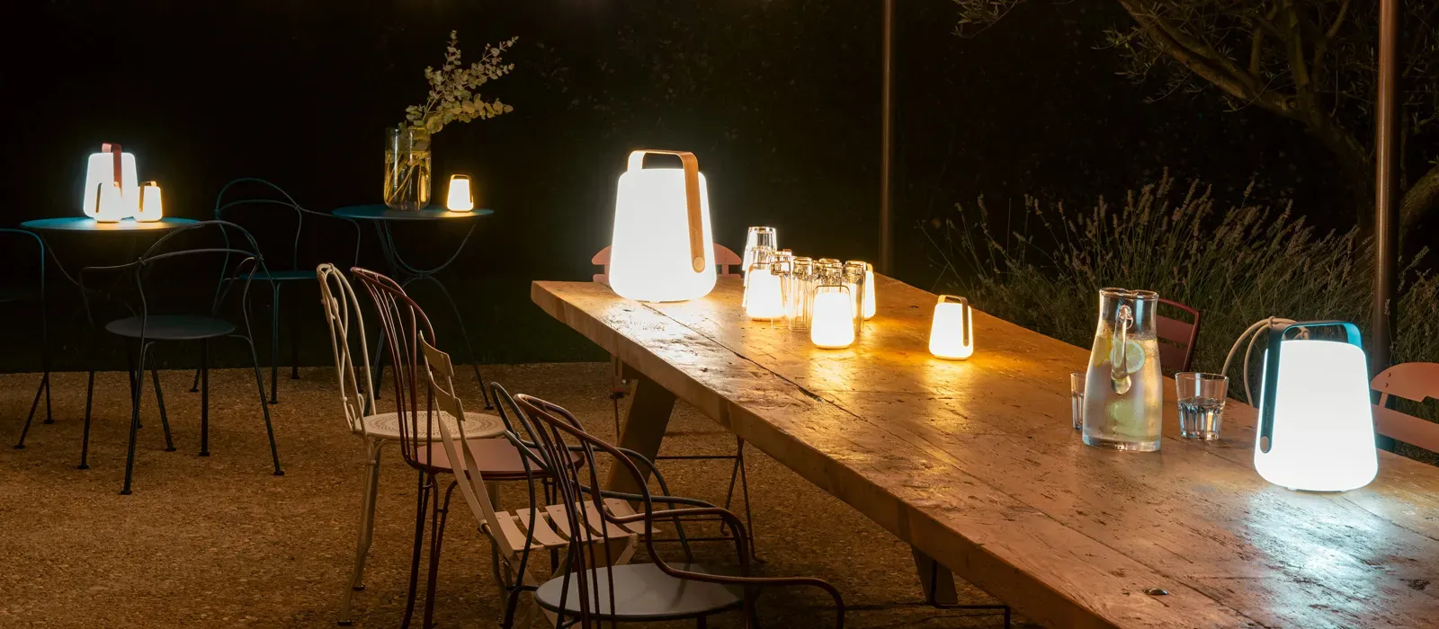 Terrace at night with a large wooden table and side tables lit by portable lamps with handles
