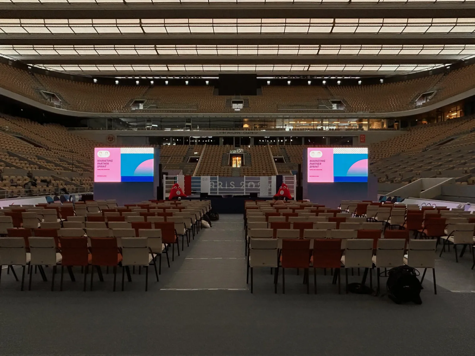 Covered court seen from the floor, with two large LED screens, a stage and rows of chairs set up