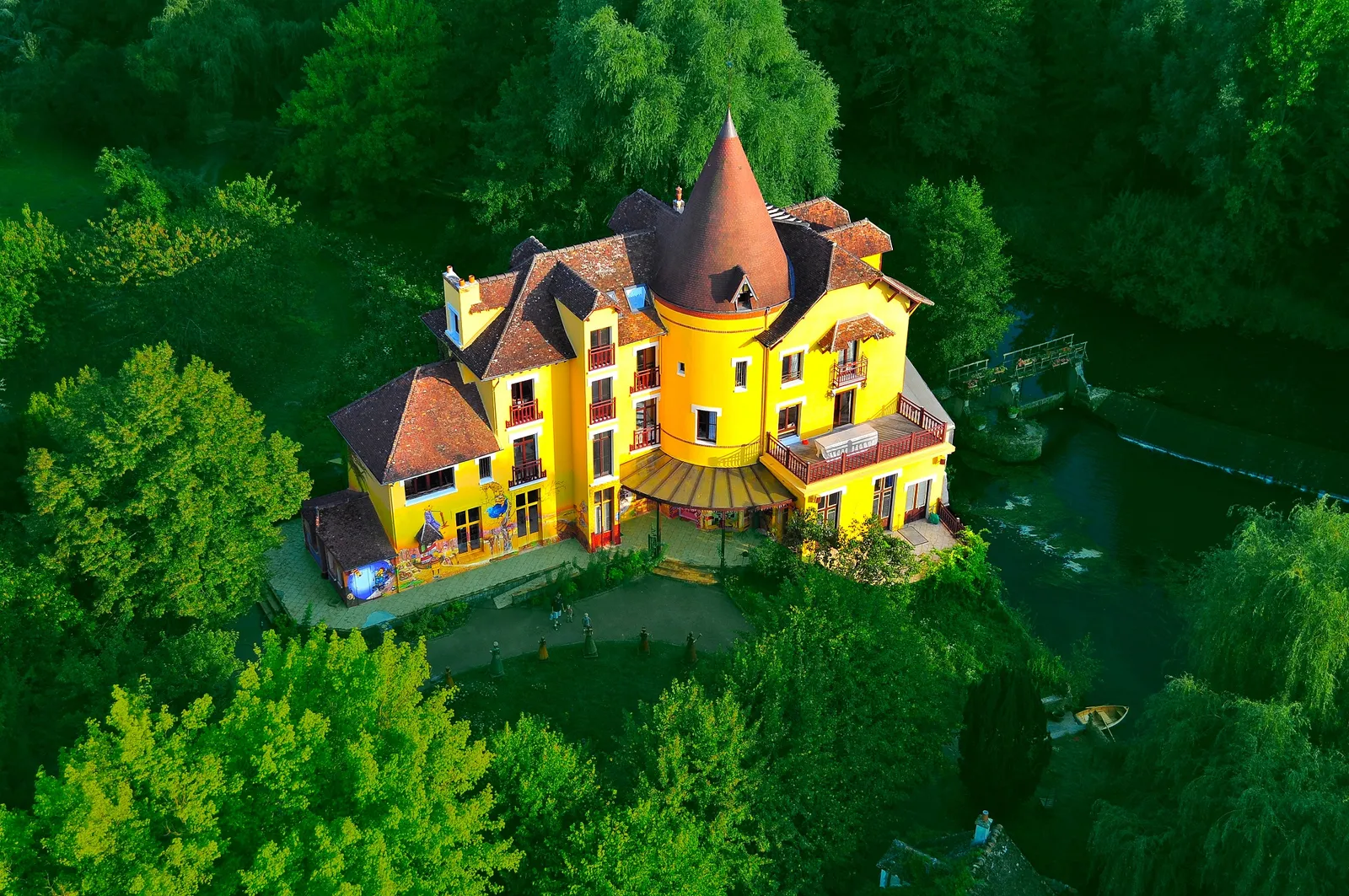 Aerial view of a yellow building with a conical turret, surrounded by trees, by the water