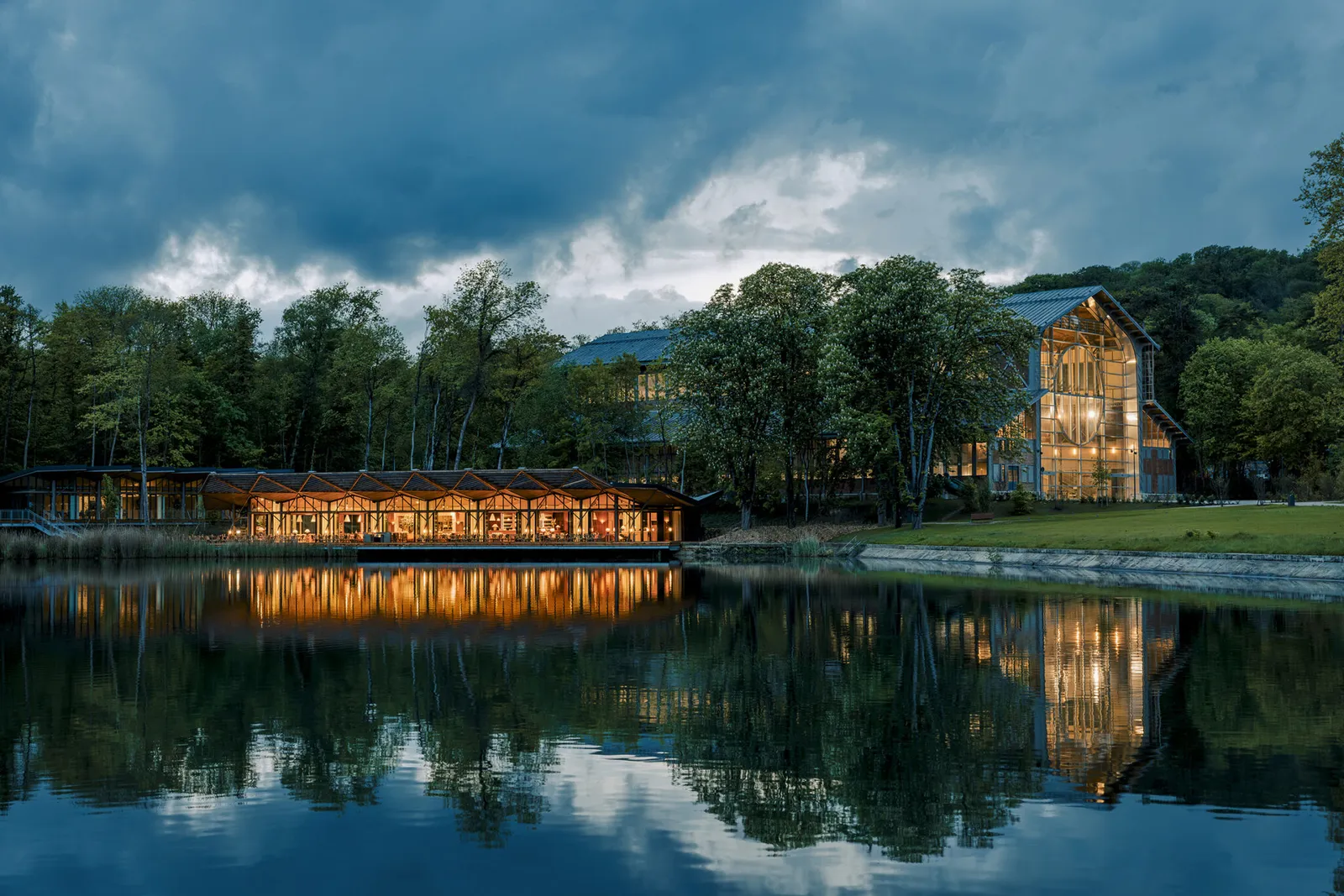 Lit glass building and wooden pavilion beside a pond at dusk